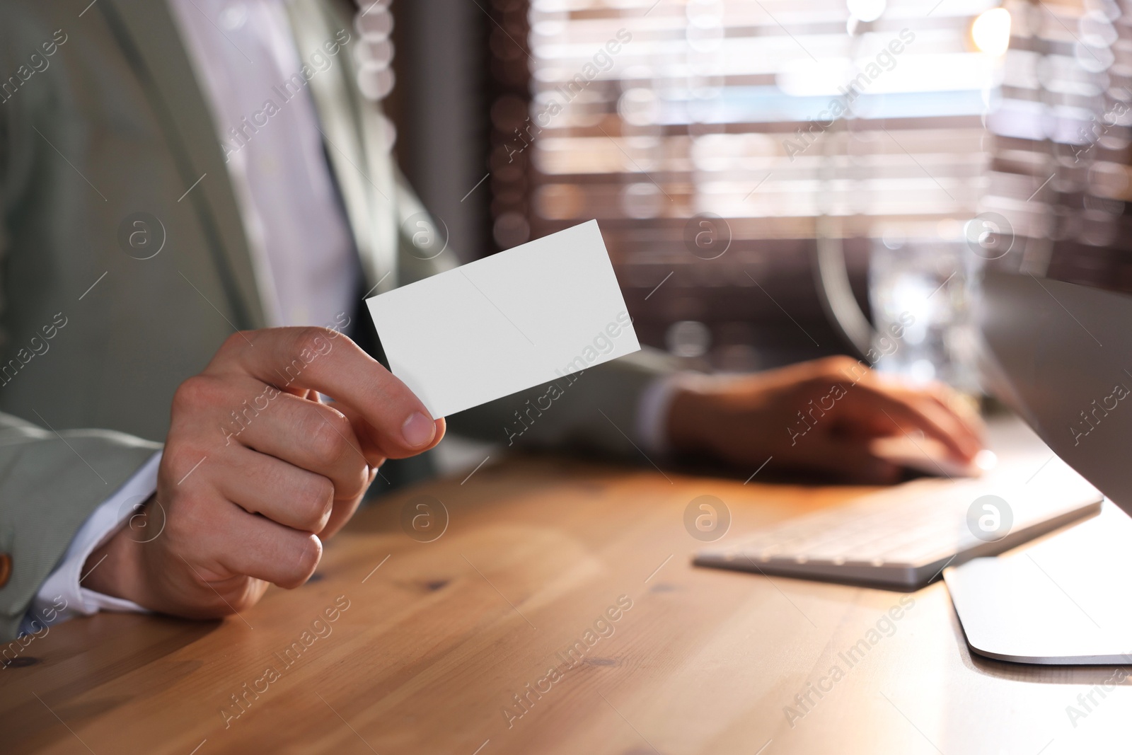 Photo of Man holding blank business card while working with computer at table in office, closeup. Mockup for design