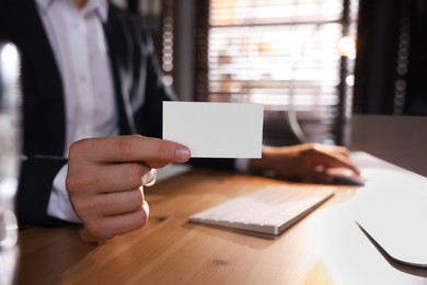 Photo of Man holding blank business card while working with computer at table in office, closeup. Mockup for design