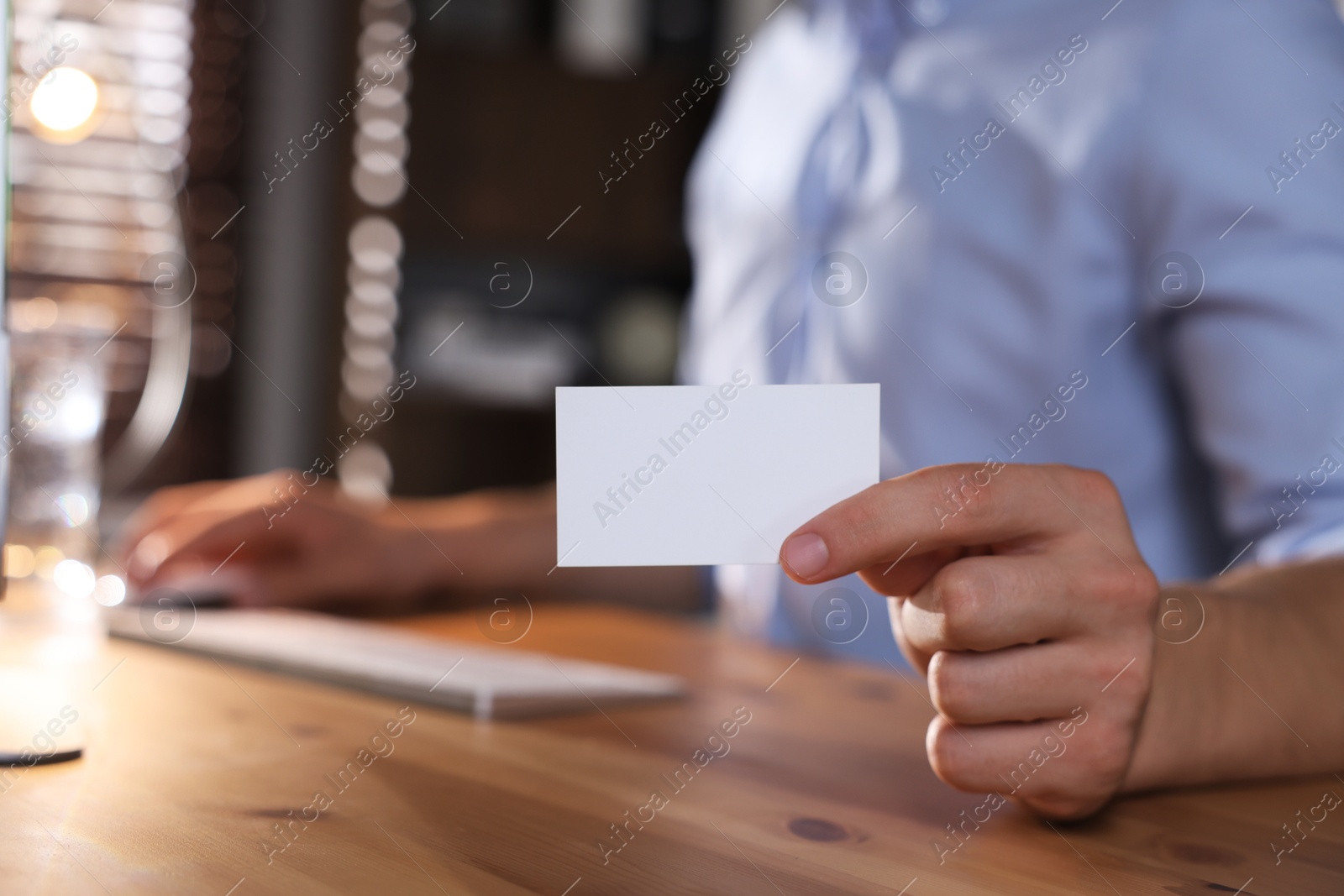 Photo of Man holding blank business card while working with computer at table in office, closeup. Mockup for design