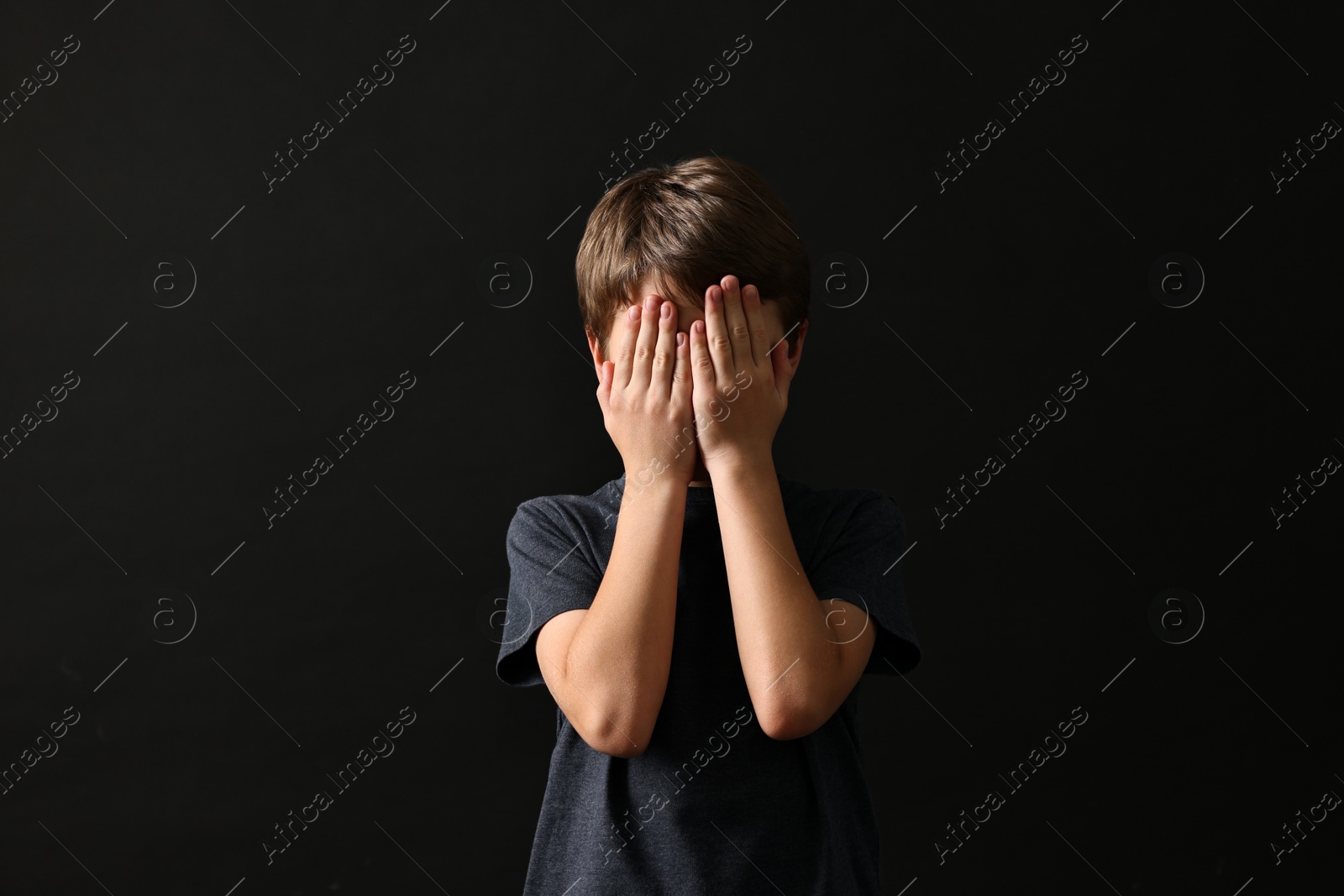 Photo of Scared little boy covering face with hands on black background