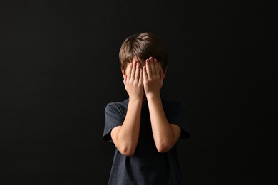 Photo of Scared little boy covering face with hands on black background