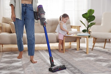 Young woman cleaning rug with cordless vacuum cleaner while her daughter drawing at home, closeup