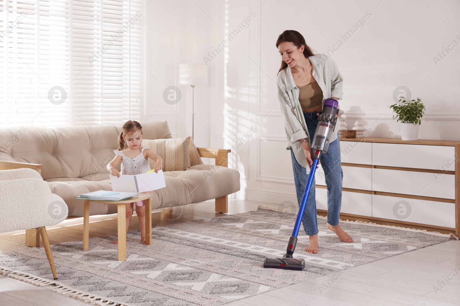 Photo of Smiling young woman cleaning rug with cordless vacuum cleaner while her daughter drawing at home