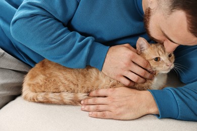 Photo of Man petting cute ginger cat on sofa at home, closeup