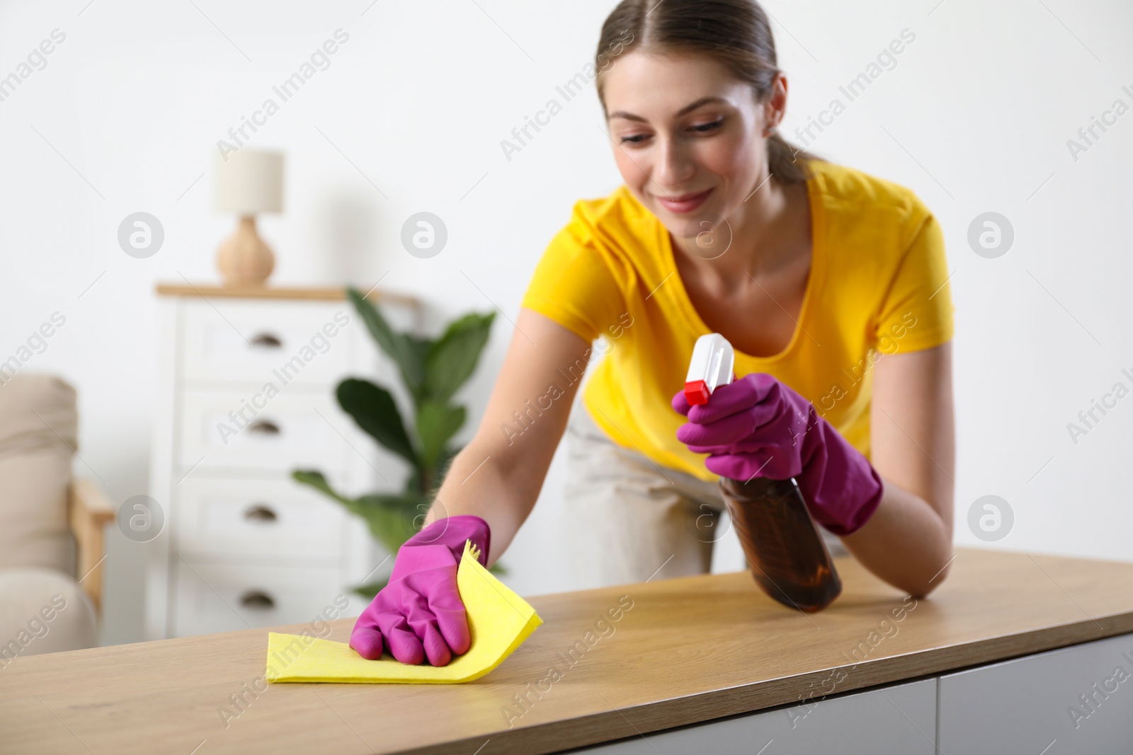 Photo of Young woman cleaning table with rag and spray at home