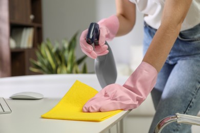 Photo of Young woman cleaning table with rag and spray in office, closeup