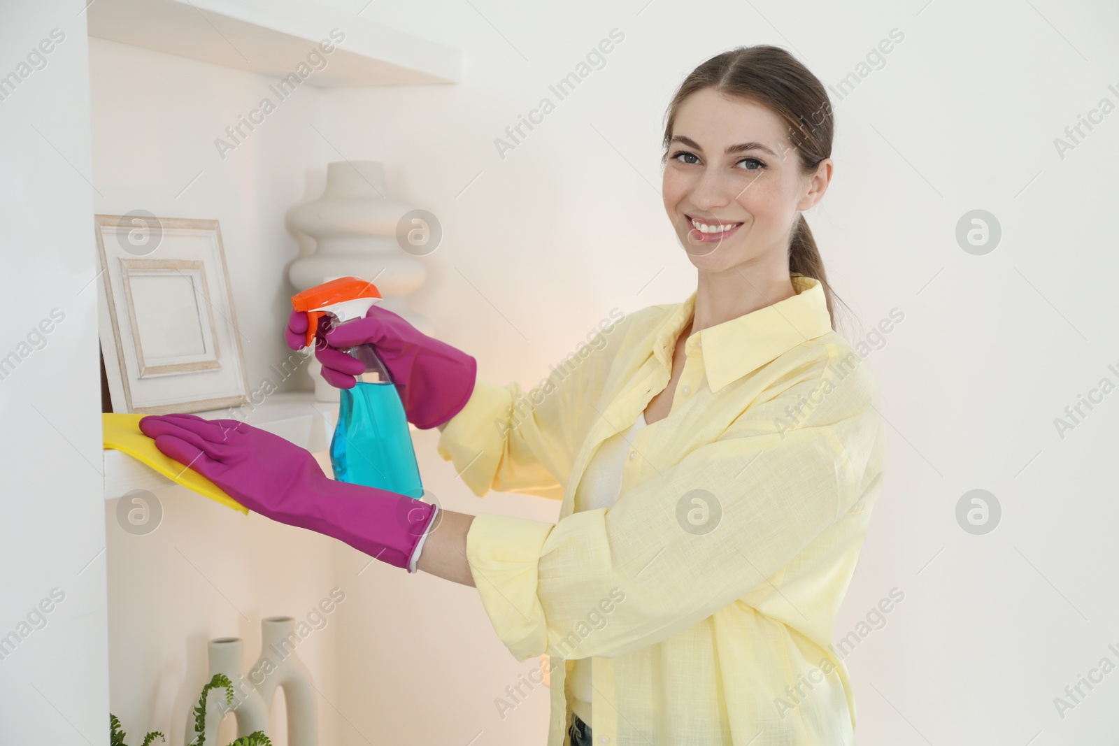 Photo of Young woman cleaning shelf with rag and spray at home