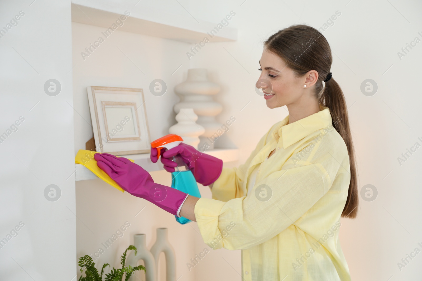 Photo of Young woman cleaning shelf with rag and spray at home