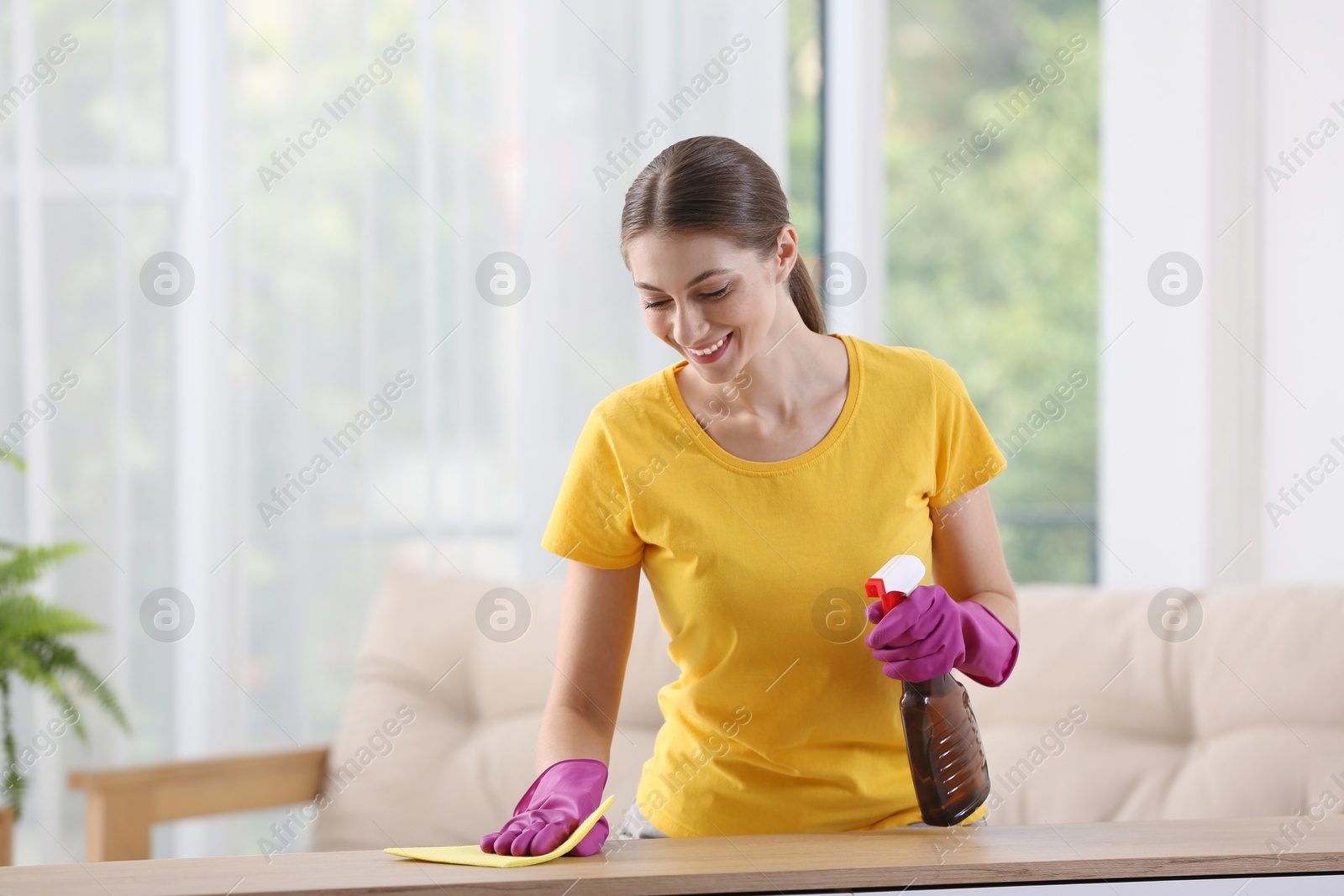 Photo of Young woman cleaning wooden table with rag and spray at home