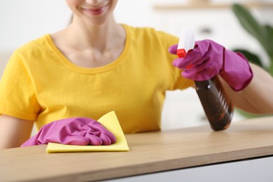 Photo of Young woman cleaning wooden table with rag and spray at home, closeup