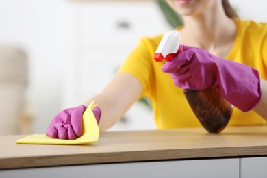 Photo of Young woman cleaning wooden table with rag and spray at home, closeup