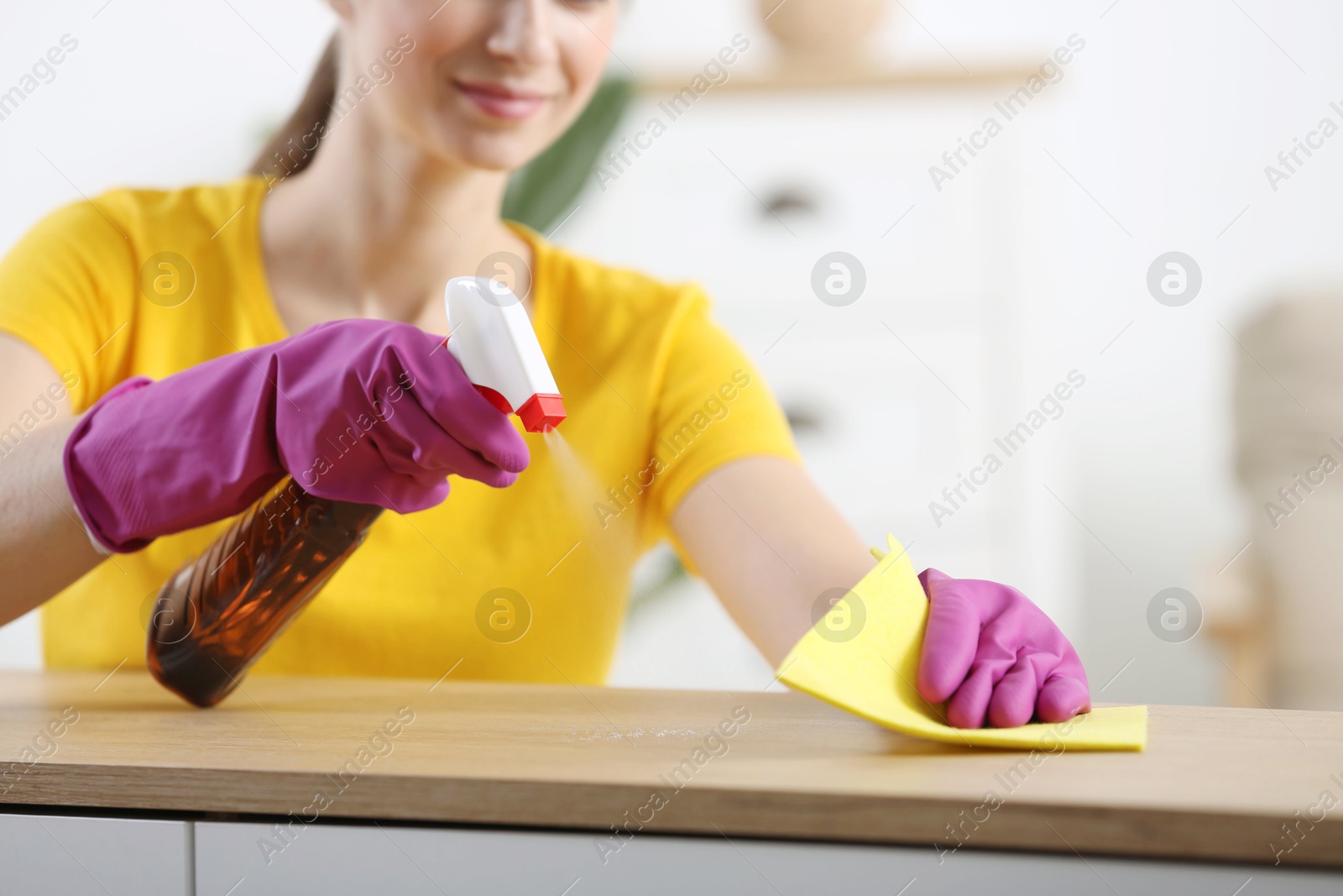 Photo of Young woman cleaning wooden table with rag and spray at home, closeup