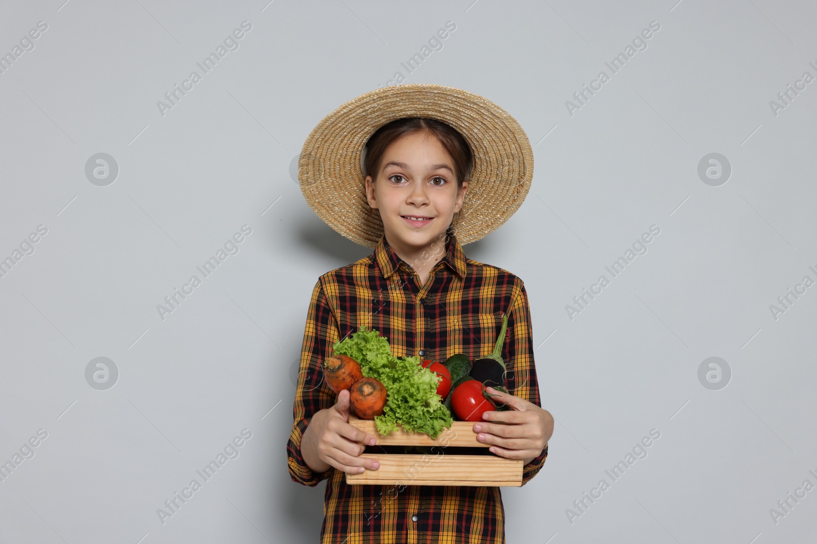 Photo of Girl with straw hat, wooden crate and vegetables pretending to be farmer on light grey background. Dreaming of future profession