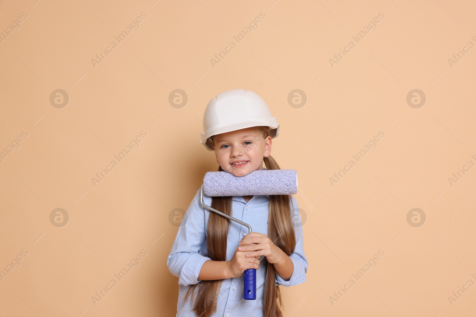 Photo of Little girl in hard hat with roller brush on beige background. Dreaming about future profession