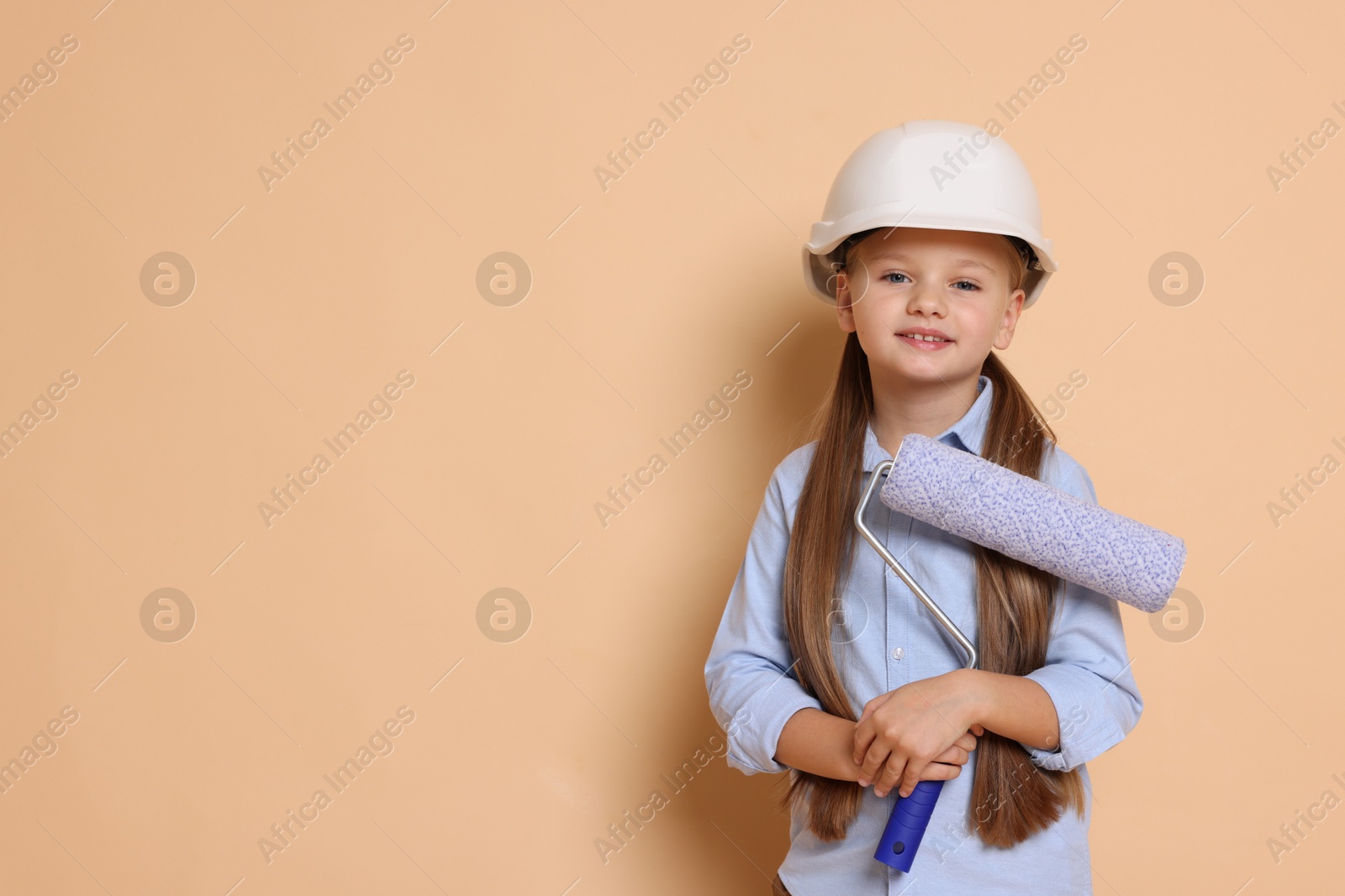 Photo of Little girl in hard hat with roller brush on beige background, space for text. Dreaming about future profession