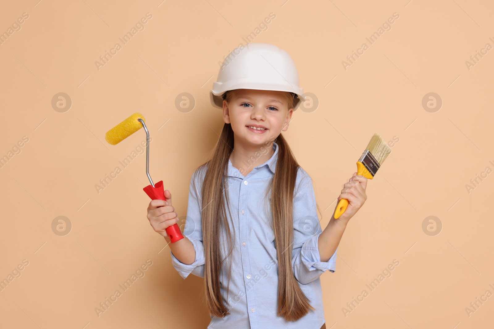 Photo of Little girl in hard hat with painting tools on beige background. Dreaming about future profession