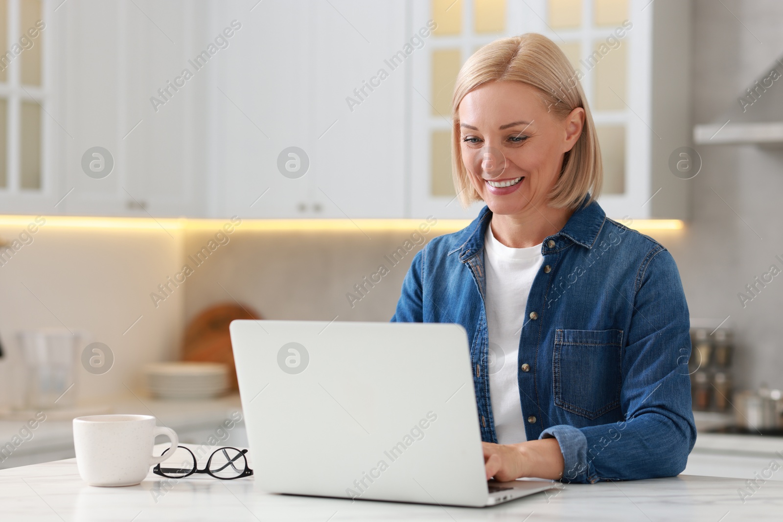 Photo of Happy middle aged woman using laptop at white marble table in kitchen