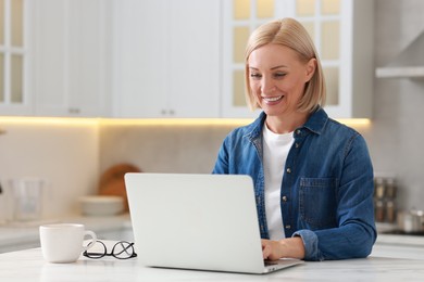 Photo of Happy middle aged woman using laptop at white marble table in kitchen