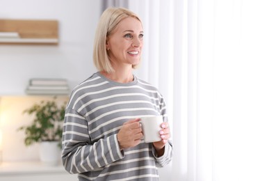 Smiling middle aged woman with cup of hot drink near window at home