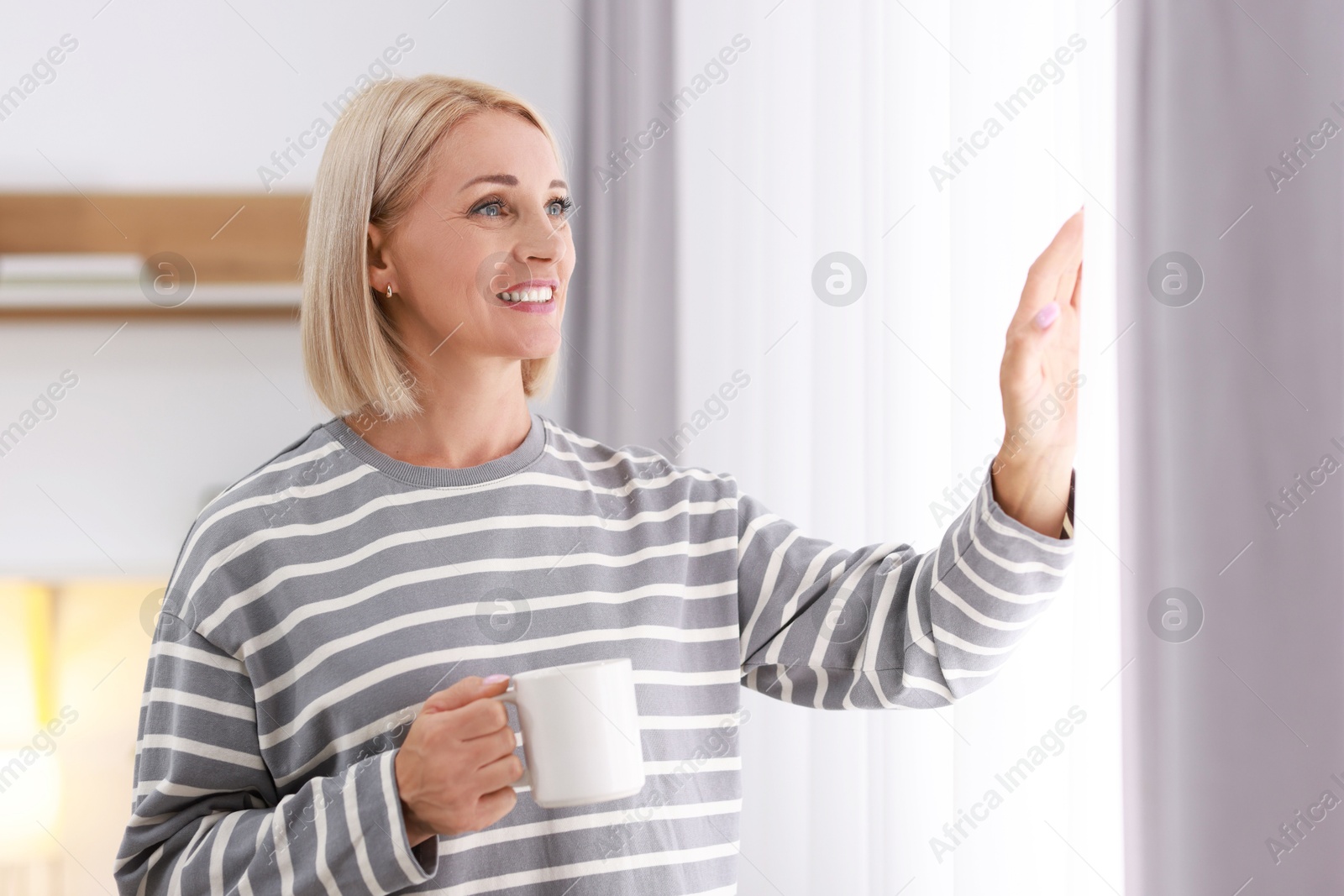 Photo of Smiling middle aged woman with cup of hot drink near window at home