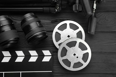 Photo of Movie clapper, camera lens and film reels on black wooden table, flat lay