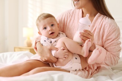 Mother holding cute little baby and bottle of milk on bed at home, closeup