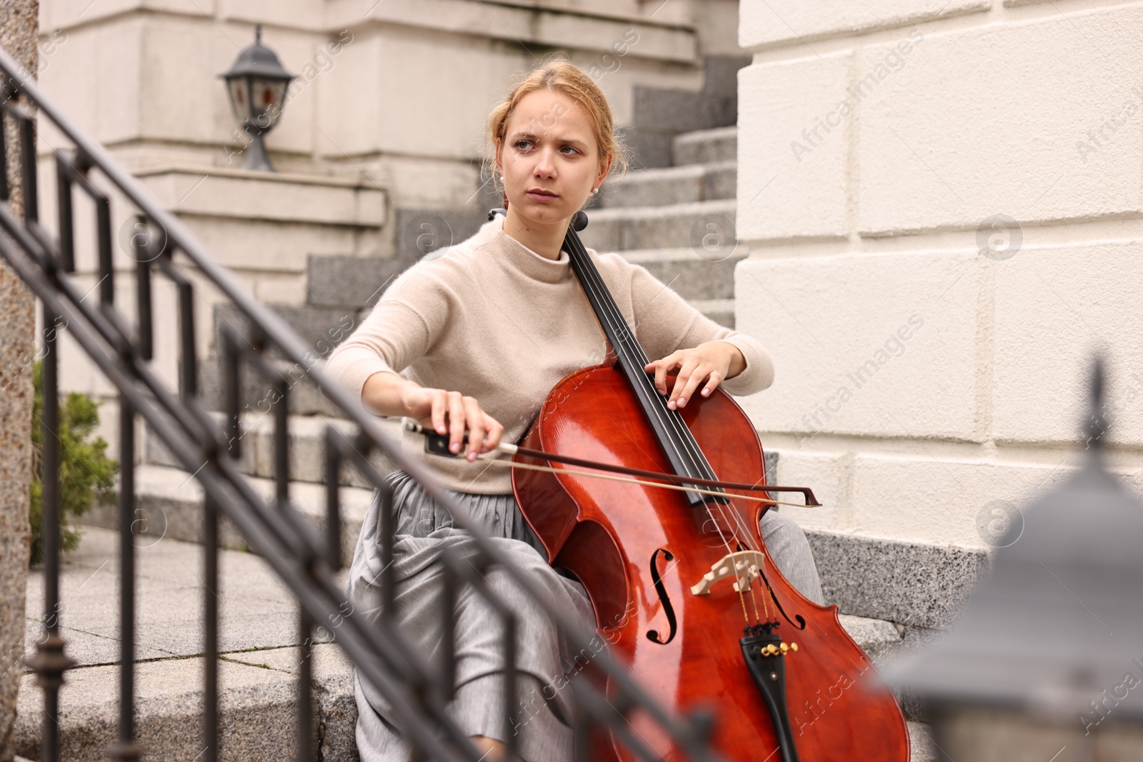 Photo of Beautiful young woman playing cello on stairs outdoors. Classic musical instrument