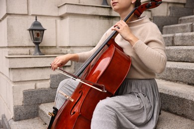 Photo of Young woman playing cello on stairs outdoors, closeup. Classic musical instrument