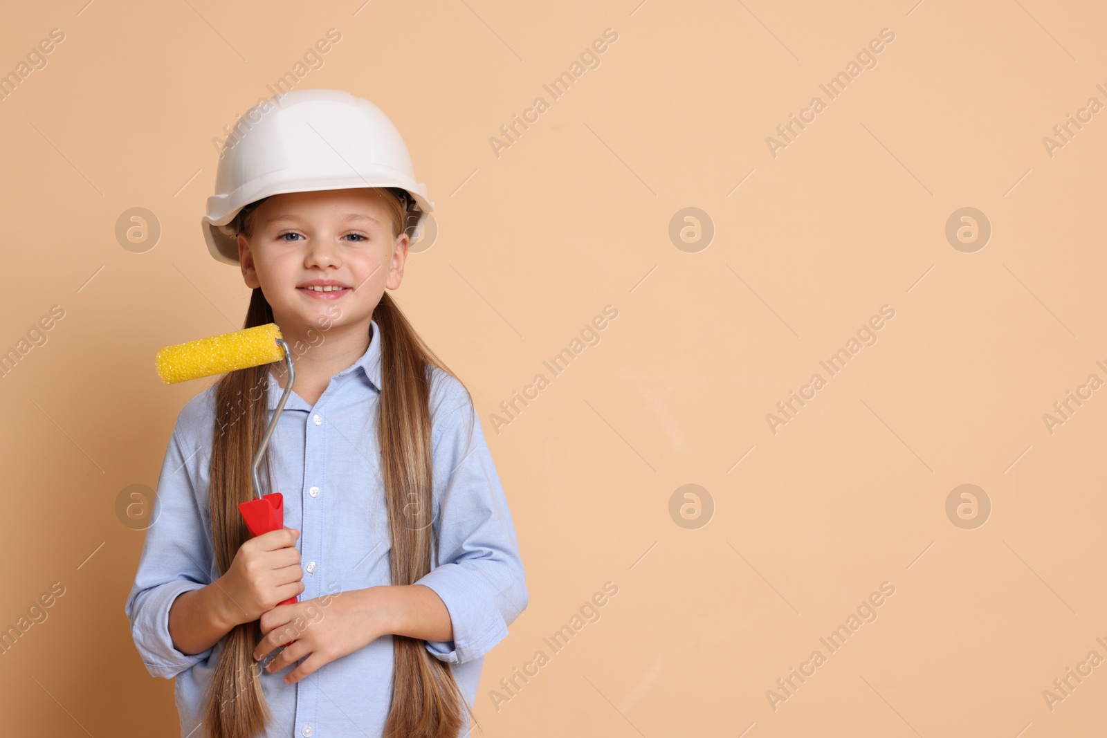 Photo of Little girl in hard hat with roller brush on beige background, space for text. Dreaming about future profession