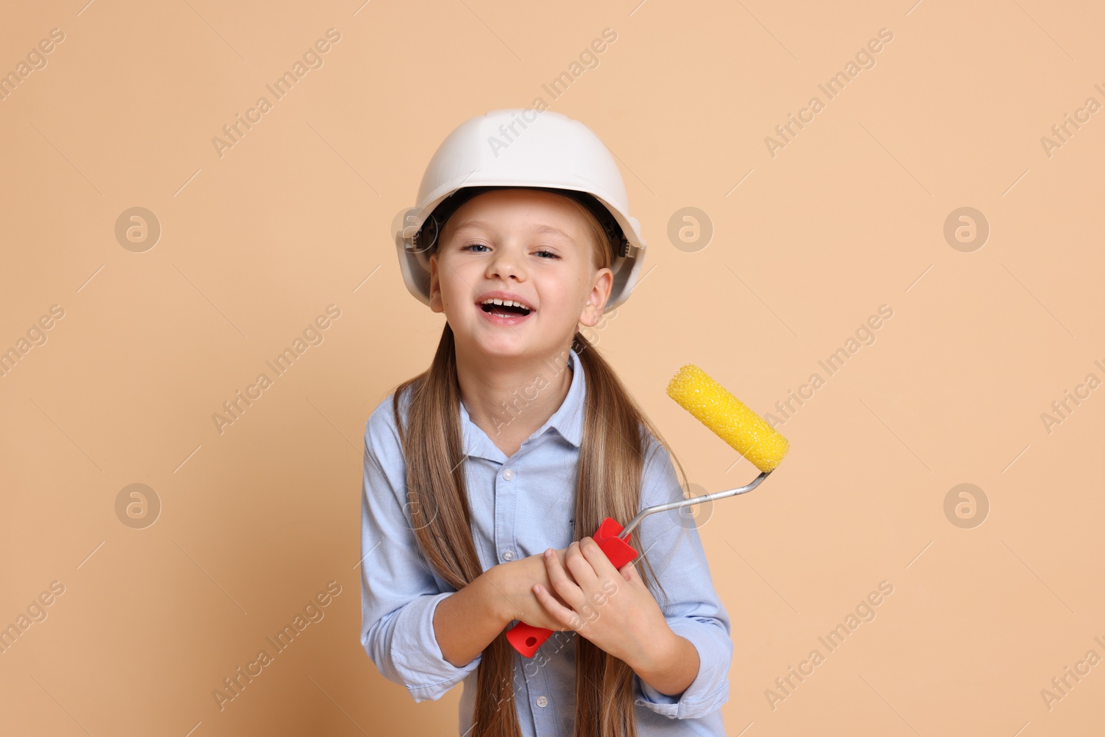 Photo of Little girl in hard hat with roller brush on beige background. Dreaming about future profession