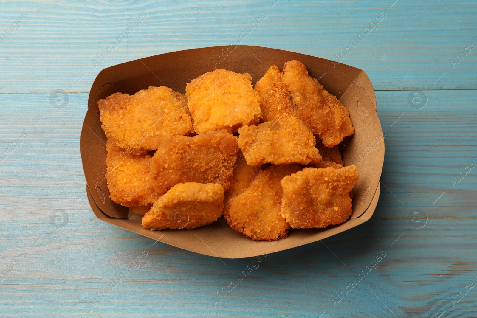 Photo of Delicious chicken nuggets in carton box on blue wooden table, top view