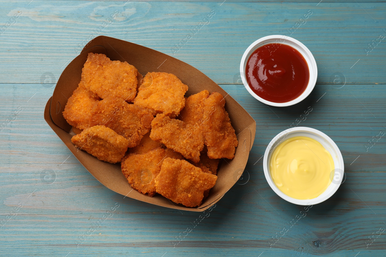 Photo of Delicious chicken nuggets in carton box with sauces on blue wooden table, flat lay