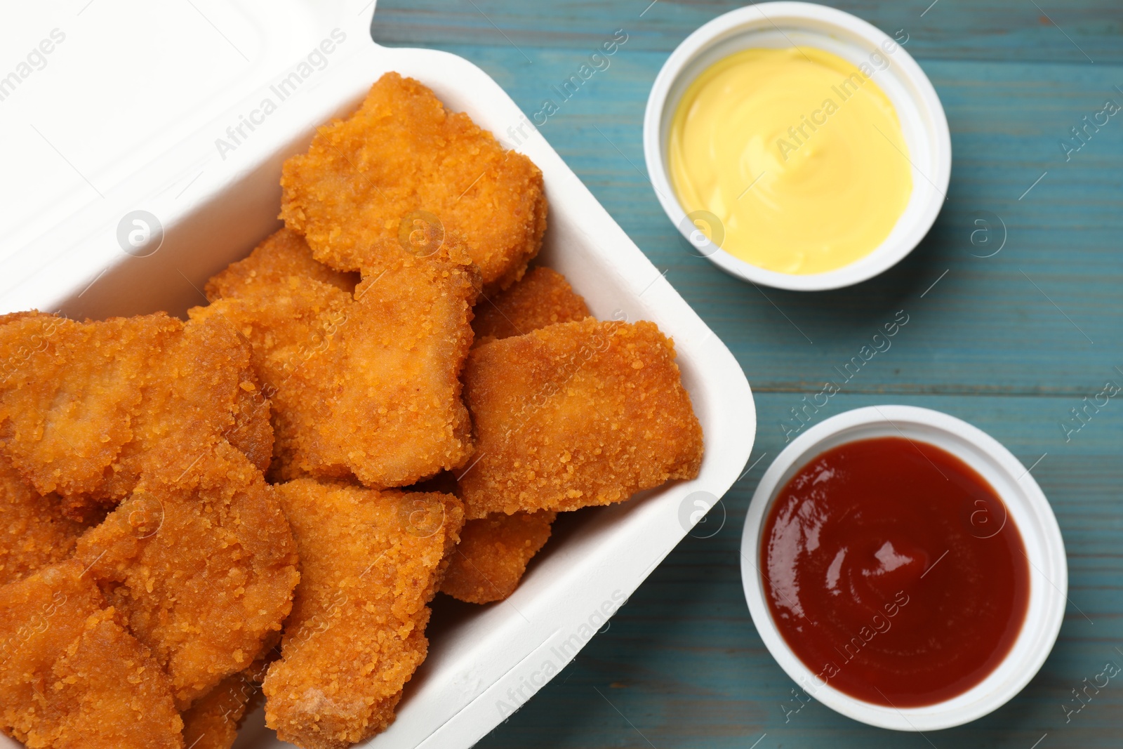 Photo of Delicious chicken nuggets in takeaway container with sauces on blue wooden table, flat lay