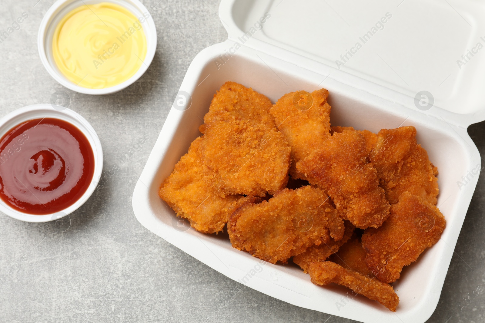 Photo of Delicious chicken nuggets in takeaway container with sauces on grey textured table, flat lay