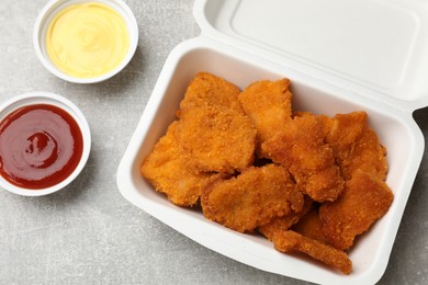 Delicious chicken nuggets in takeaway container with sauces on grey textured table, flat lay