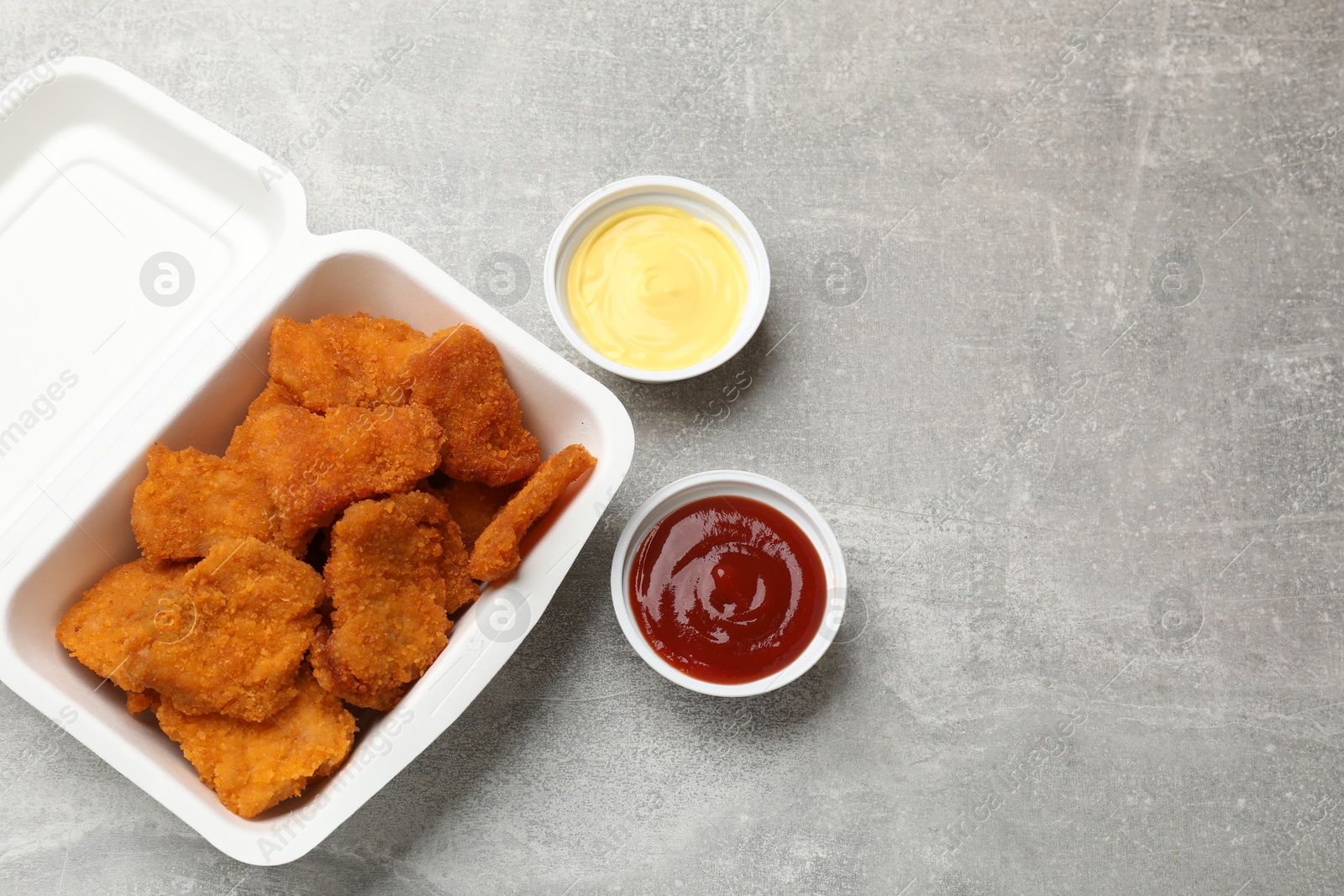 Photo of Delicious chicken nuggets in takeaway container with sauces on grey textured table, flat lay. Space for text