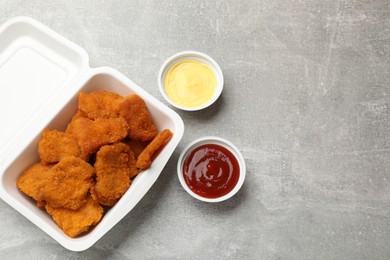 Photo of Delicious chicken nuggets in takeaway container with sauces on grey textured table, flat lay. Space for text