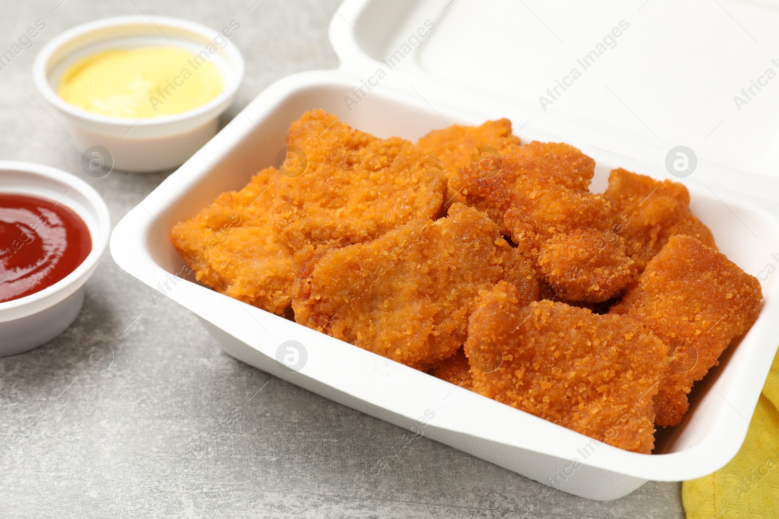 Photo of Delicious chicken nuggets in takeaway container with sauces on grey textured table, closeup