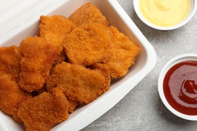 Photo of Delicious chicken nuggets in takeaway container with sauces on grey textured table, flat lay