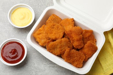 Photo of Delicious chicken nuggets in takeaway container with sauces on grey textured table, flat lay