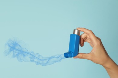 Asthma treatment. Woman holding inhaler on light blue background, closeup. Aerosolized medication coming out of device