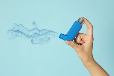 Asthma treatment. Woman holding inhaler on light blue background, closeup. Aerosolized medication coming out of device