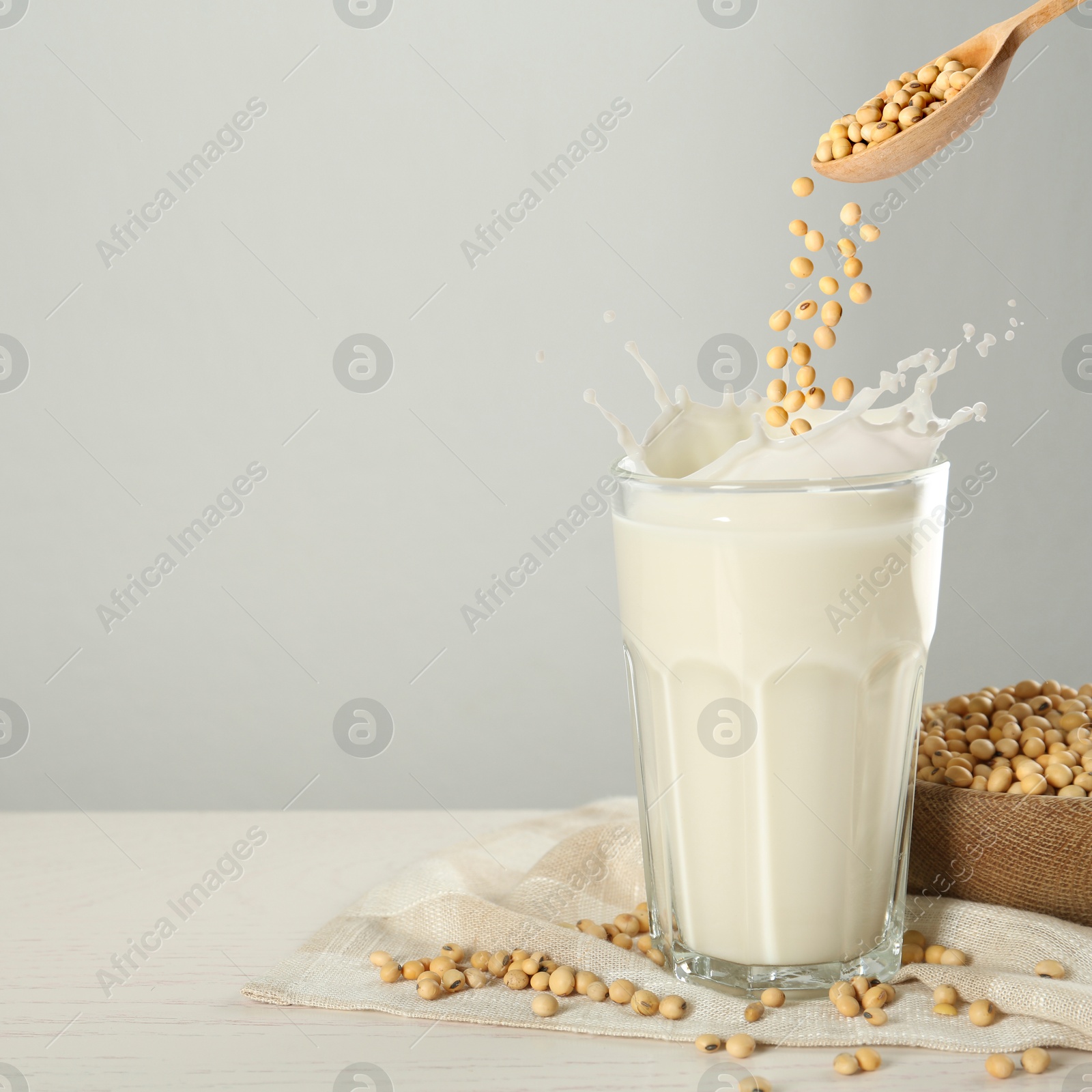 Image of Soy beans falling from wooden spoon into glass of plant milk on white table. Space for text