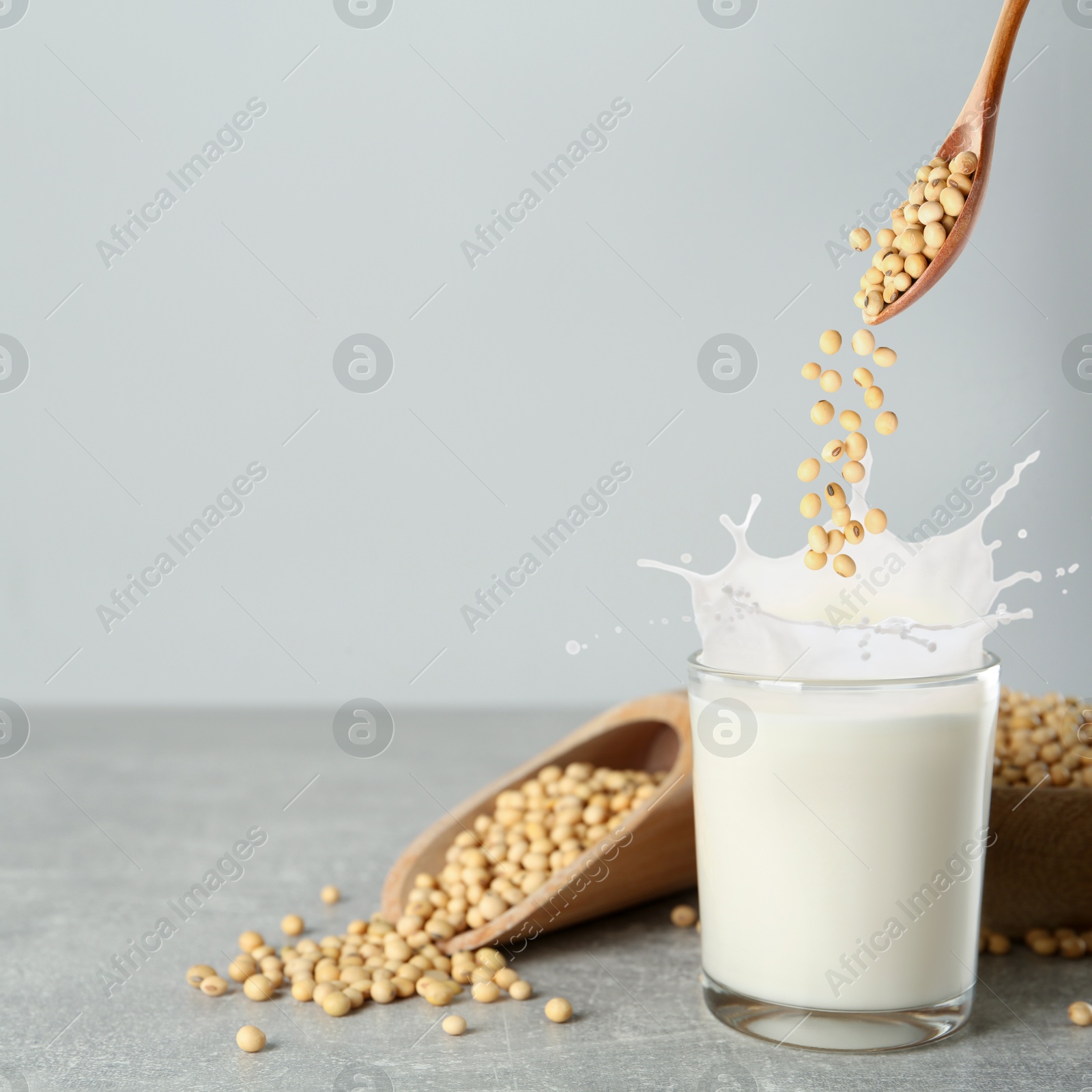 Image of Soy beans falling from wooden spoon into glass of plant milk on grey table. Space for text