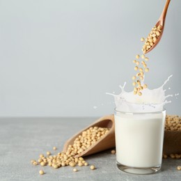 Image of Soy beans falling from wooden spoon into glass of plant milk on grey table. Space for text