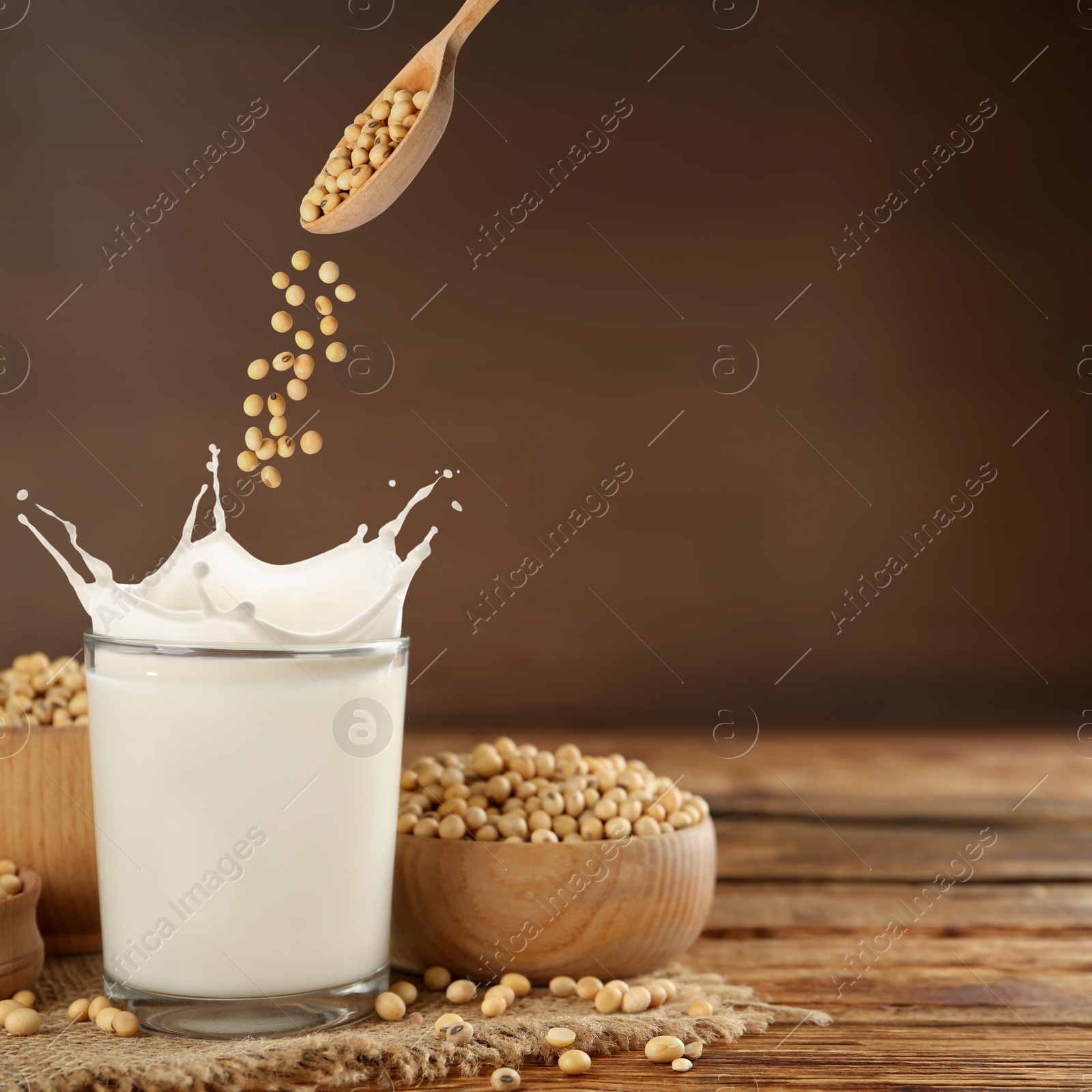 Image of Soy beans falling from spoon into glass of plant milk on wooden table. Space for text