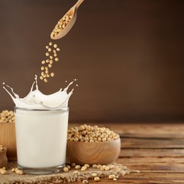 Image of Soy beans falling from spoon into glass of plant milk on wooden table. Space for text