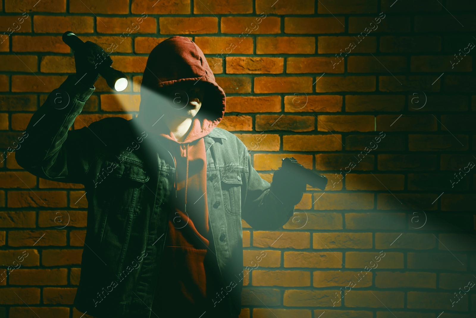 Image of Thief in hoodie with gun and flashlight against red brick wall. Armed robbery