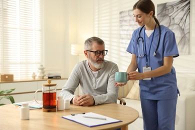 Photo of Caregiver giving cup of drink to senior man indoors. Home health care service