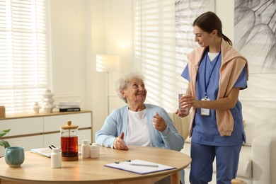 Caregiver giving glass of water to senior woman indoors. Home health care service